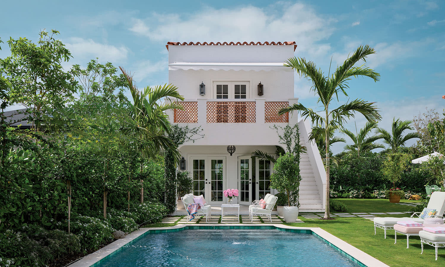 Elegant villa with white facade and terracotta tiles, lush garden, palm trees, and a serene blue pool. Cozy chairs surround the tropical oasis.